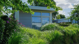 Gemütliche Pavilion-Lodge mit großen Fenstern, umgeben von grünen Büschen und Blumen an einem sonnigen Tag.