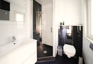 Moderne badeværelse på hytte med væghængt toilet, bruseniche, hvid vask og sorte fliser ved vindue.
