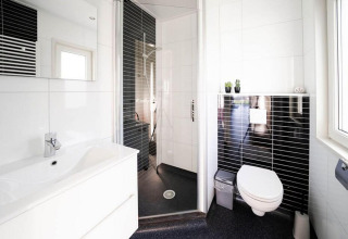 Baño moderno en una cabaña con inodoro suspendido, ducha acristalada, lavabo blanco y azulejos negros.