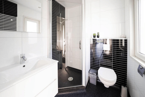 Moderne badeværelse på hytte med væghængt toilet, bruseniche, hvid vask og sorte fliser ved vindue.