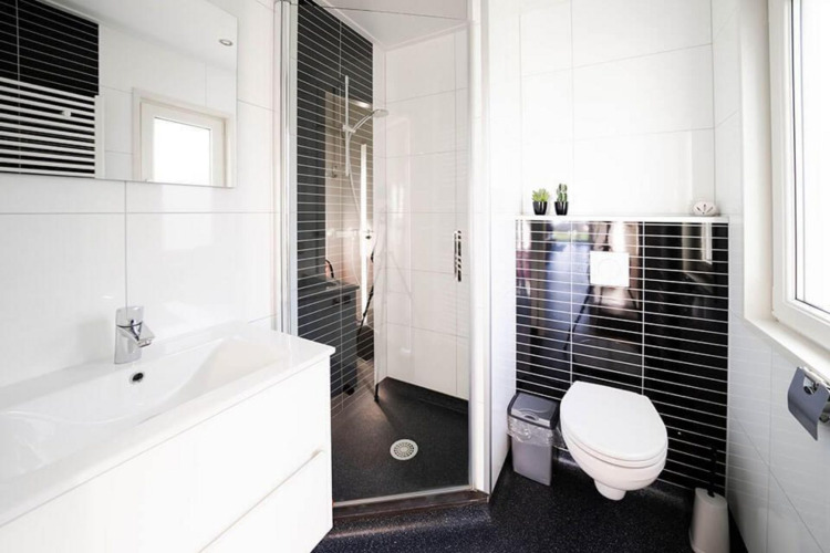 Baño moderno en una cabaña con inodoro suspendido, ducha acristalada, lavabo blanco y azulejos negros.