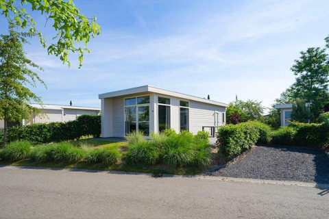Modernes Ferienhaus mit großen Fenstern, umgeben von grünen Sträuchern, bei sonnigem Wetter.
