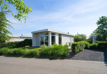 Moderne lodge met grote ramen, omringd door groen en struiken, onder een heldere blauwe hemel.