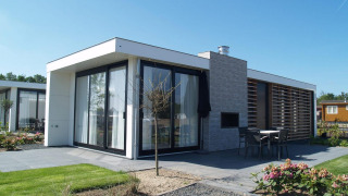 Moderne paviljoenlodge met grote ramen, terras en tuin onder een blauwe hemel, ideaal voor ontspanning.