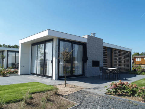 Moderne pavillon-lodge med store vinduer, terrasse og have under en blå himmel, perfekt til afslapning.
