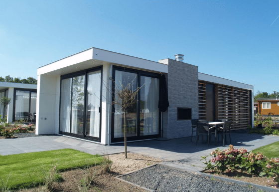 Moderne Pavillon-Lodge mit großen Fenstern, Terrasse und Garten unter blauem Himmel, ideal zum Entspannen.