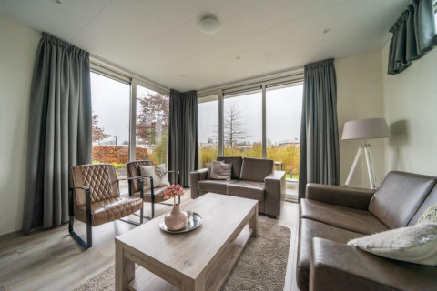 Salon moderne avec grandes fenêtres, canapés en cuir et rideaux gris au Pavilion lodge d’EuroParcs Veluwemeer.
