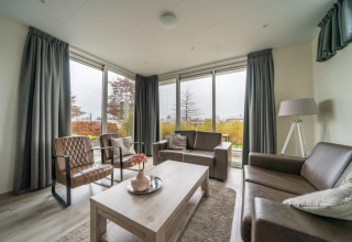 Salon moderne avec grandes fenêtres, canapés en cuir et rideaux gris au Pavilion lodge d’EuroParcs Veluwemeer.