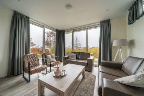 Modernes Wohnzimmer mit großen Fenstern, Lederstühlen und grauen Vorhängen in der Pavilion-Lodge EuroParcs Veluwemeer.