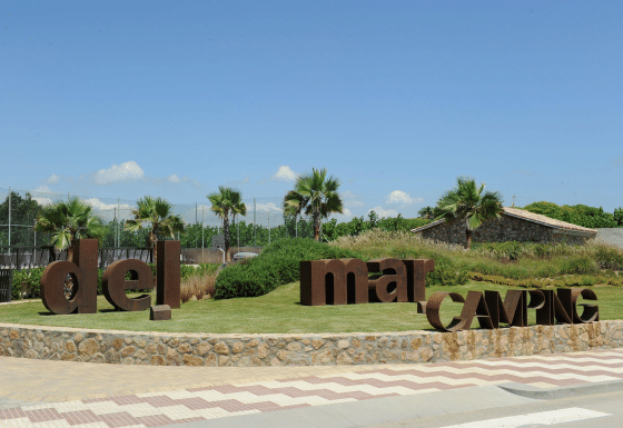 Entrada del camping Camping del Mar en Cataluña, España, con palmeras y grandes letras marrones en el césped.