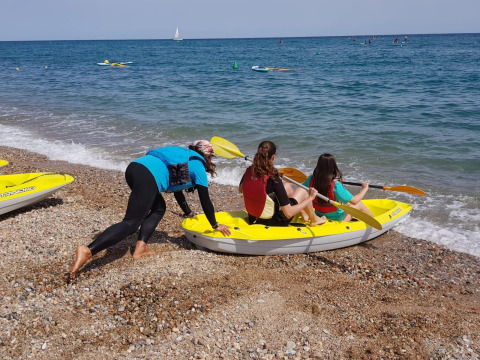 Three people prepare to launch a yellow kayak from a pebbly beach at Camping del Mar in Catalonia, Spain.