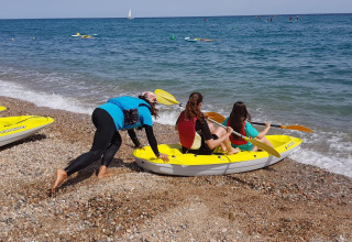 Three people prepare to launch a yellow kayak from a pebbly beach at Camping del Mar in Catalonia, Spain.