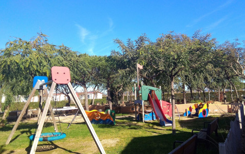 Parque infantil en Camping del Mar en Cataluña, España con columpios, tobogán, bancos y árboles verdes.