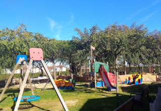 Playground at Camping del Mar holiday park in Catalonia, Spain with swings, slide, benches and green trees.