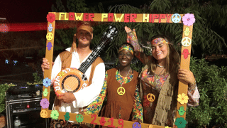 Tres personas disfrazadas de hippies posan con un marco de Flower Power en Camping del Mar, España.