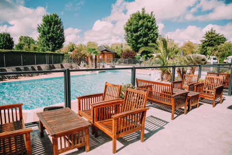 Área de piscina al aire libre con asientos de madera y palmeras en Camping Seasonova Haliotis en Normandía, Francia.