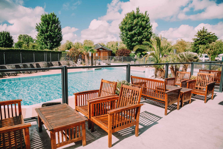 Outdoor pool area with wooden seating and palm trees at Camping Seasonova Haliotis holiday park in Normandy, France.