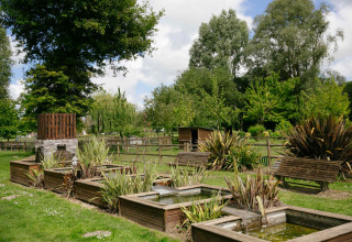Grüner Ferienpark mit Holzbänken, Pflanzen und kleinen Teichen bei Camping Seasonova Haliotis in der Normandie.