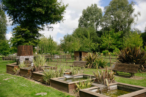 Groen vakantiepark met houten zitbanken, beplanting en kleine vijvertjes bij Camping Seasonova Haliotis in Normandië.