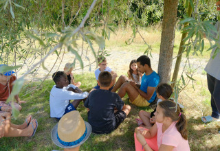 Kinder und ein Erwachsener sitzen im Kreis im Schatten auf Camping Seasonova Haliotis in der Normandie.