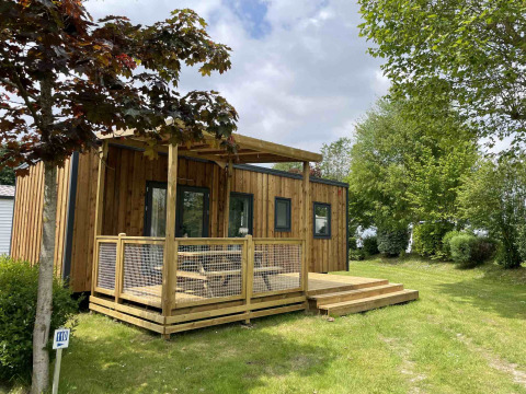 Cabaña de madera con terraza rodeada de vegetación en Camping Seasonova Haliotis, Normandía, Francia.