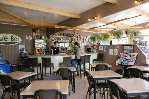 Vista interior del café bar en Camping Seasonova Haliotis con mesas, sillas y plantas colgantes.