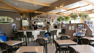 Vista interior del café bar en Camping Seasonova Haliotis con mesas, sillas y plantas colgantes.