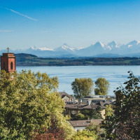 Vista de Les Brenets, Neuchâtel, Suiza, con una torre de iglesia, lago y los Alpes nevados al fondo.