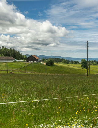 Paisaje rural cerca de Les Brenets, Neuchâtel, Suiza, con prados verdes, granjas y cielo parcialmente nublado.