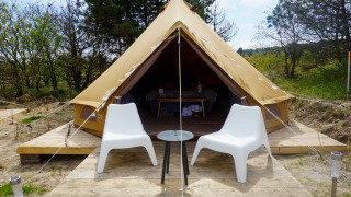 En Bell Escape Family tipi-telt med to hvide stole og et lille bord udenfor, omgivet af natur og træer.