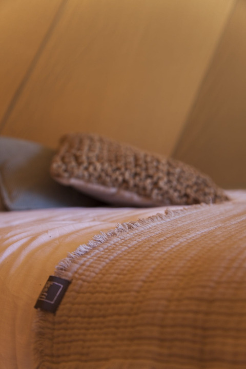 Primer plano de una cama con manta beige y cojín dentro de una tienda tipi iluminada con luz cálida.