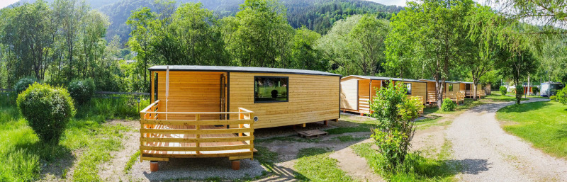 Chalets en bois à Drau Camping Sachsenburg, Carinthie, Autriche, entourés de verdure et de montagnes.