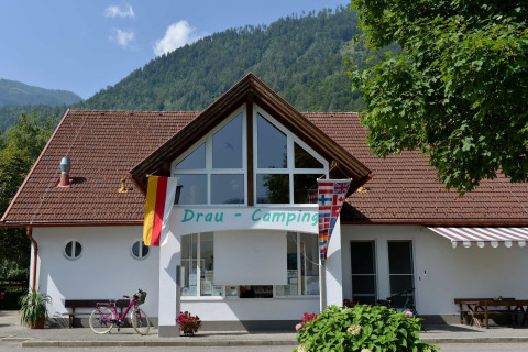 Eingang zum Drau Camping Sachsenburg, mit Fahnen, Bergen und Fahrrad in Kärnten, Österreich.