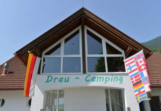 Indgang til Drau Camping Sachsenburg i Østrig, med vinduer, tyske og internationale flag udenfor.