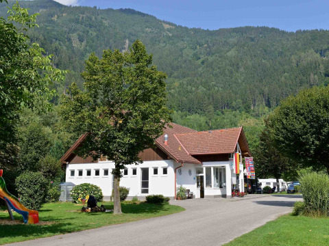 Drau Camping Sachsenburg i Kärnten, Østrig, med et hovedhus, grønne træer og bjerge i baggrunden.
