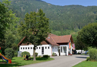 Drau Camping Sachsenburg in Karinthië, Oostenrijk, met het hoofdgebouw, bomen en groene heuvels op de achtergrond.