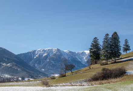 Paisaje en los alrededores de Kramsach, Tirol, Austria, con montañas nevadas, colinas verdes y pinos bajo cielo azul.