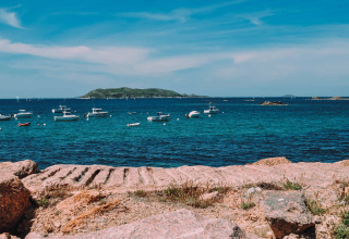 Både flyder på blåt hav ved Camping Seasonova Les 7 Iles i Bretagne, Frankrig, med ø i baggrunden.