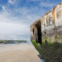 Foto de una antigua estructura de hormigón en la playa, cubierta de algas, con la costa al fondo.