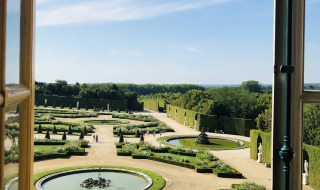Vista desde una ventana abierta a los elegantes jardines formales de Versalles bajo un brillante cielo azul.
