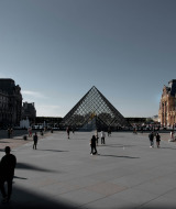 Foto de la Pirámide del Louvre y la fachada del museo en París, con visitantes caminando por la explanada.