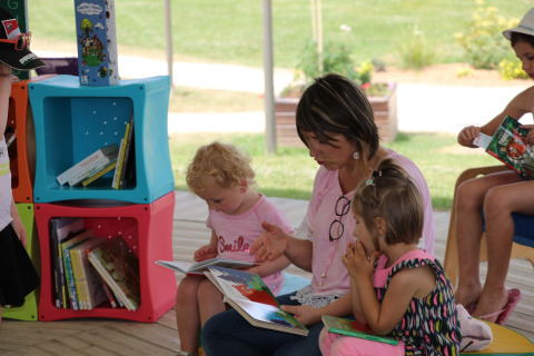 An adult reads a picture book to two children in an outdoor library area at Camping Seasonova L'Etang des Bois.