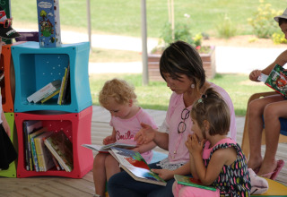 Een volwassene leest een prentenboek voor aan twee kinderen in een buitenbibliotheek bij Camping Seasonova L'Etang des Bois.