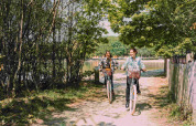To personer cykler på en solrig grussti omgivet af træer i Camping Seasonova L'Etang des Bois, Frankrig.