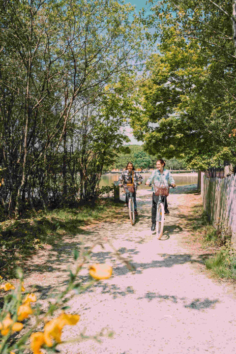 Two people cycling on a sunny path surrounded by trees at Camping Seasonova L'Etang des Bois, France.