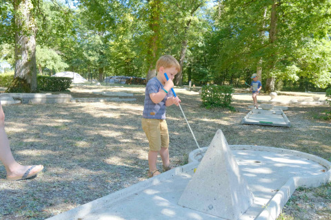 Zwei Kinder spielen Minigolf im Freien zwischen schattigen Bäumen im Urlaubspark L'Etang des Bois, Frankreich.