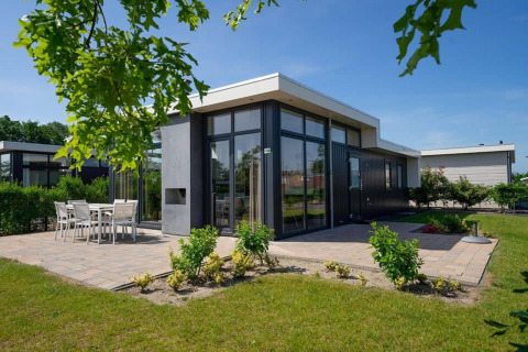 Moderne Pavilion-lodge met grote ramen, terras en tuinset onder een heldere blauwe hemel.