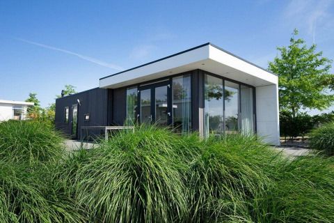 Moderne Pavilion Waterfront lodge met grote ramen en veel groen, gefotografeerd onder een blauwe lucht.