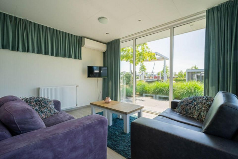 Stue i Pavilion Waterfront-lodge ved EuroParcs Veluwemeer med store vinduer, sofaer og udsigt til haven.