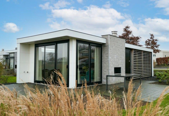 Moderne Pavilion Waterfront lodge met grote ramen, omgeven door gras en een stralend blauwe lucht.